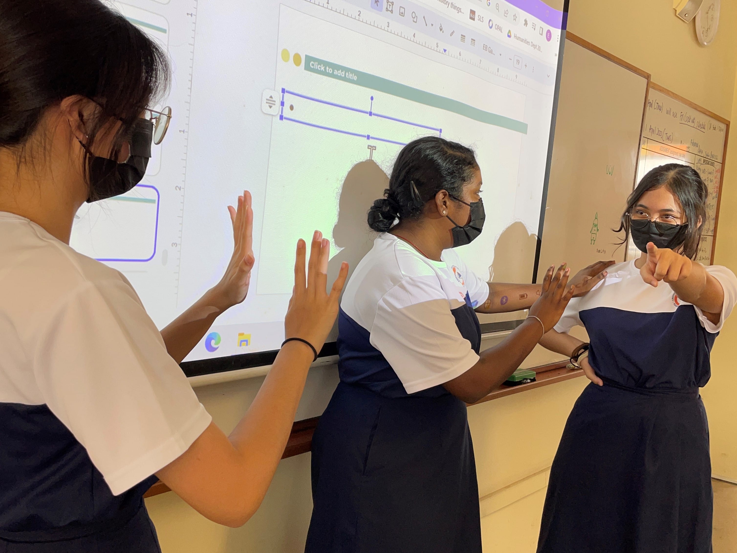 Three masked female students in uniforms stand in front of a classroom projector screen, gesturing with their hands.